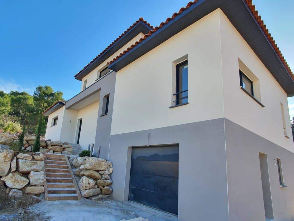 Façade moderne bicolore: Crépi gris et beige pour construction neuve. Maison moderne à façade bicolore (gris et beige) sous un ciel bleu. Toit en tuiles romanes, garage intégré, escalier en bois avec aménagement rocheux.
