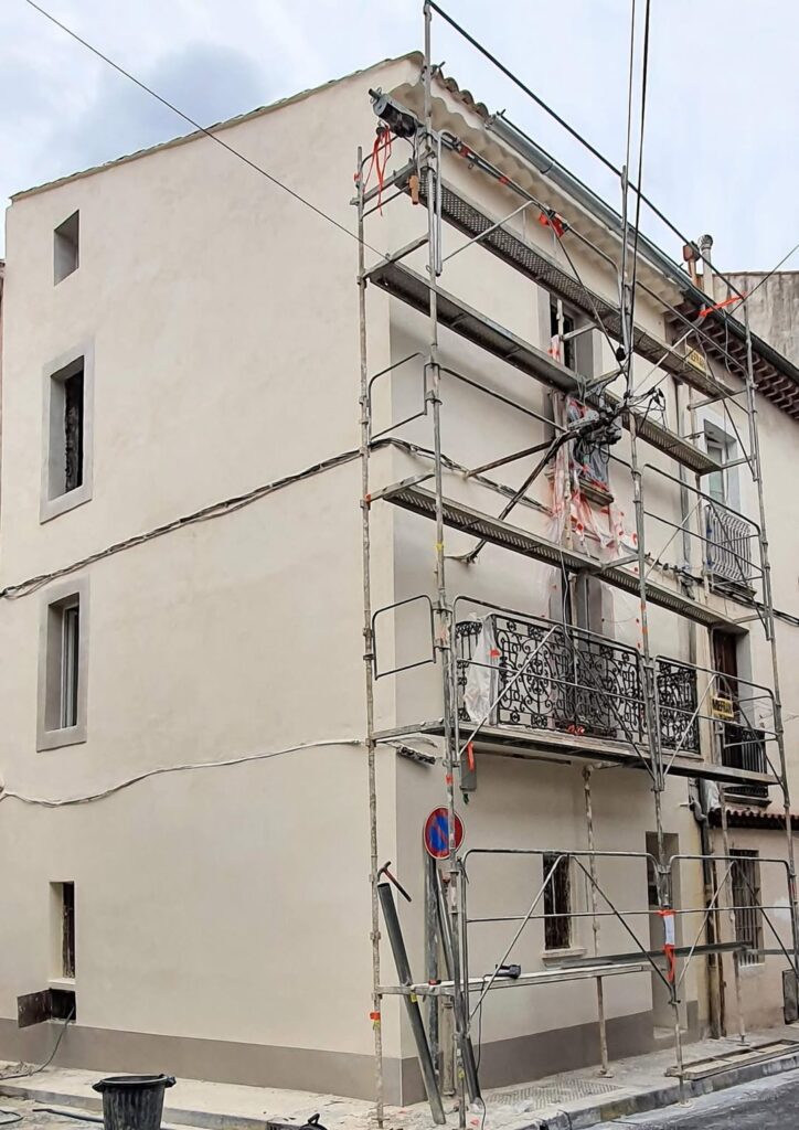 Rénovation de Façade avec Échafaudage pour Enduit Neuf Façade en cours de rénovation recouverte d'un enduit beige clair. Échafaudage métallique, treuil de chantier et balcon en fer forgé. Lignes électriques visibles.