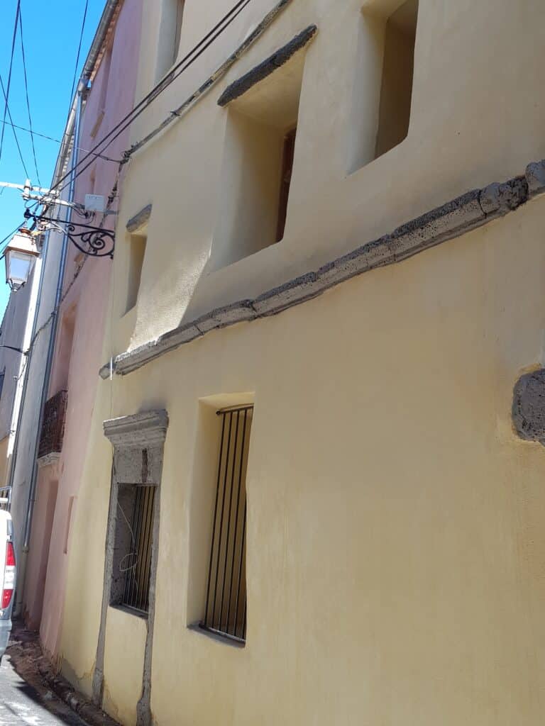 Façade ocre d'une ruelle ancienne avec fenêtres en pierre et lampadaire en fer forgé sous un ciel bleu ensoleillé.
