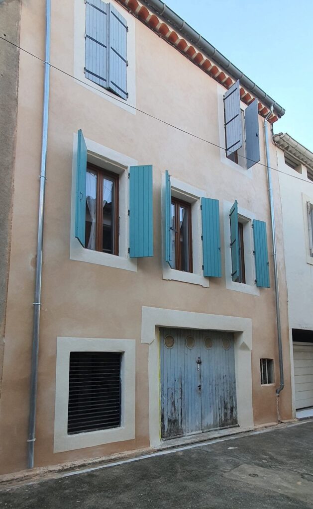 Façade ocre d'une maison traditionnelle avec volets bleus ouverts et grande porte de garage en bois vieilli.